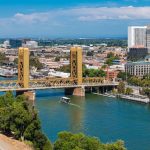 Aerial view of Sacramento, California, featuring the golden Tower Bridge over the Sacramento River, a boat, urban buildings, and the State Capitol.