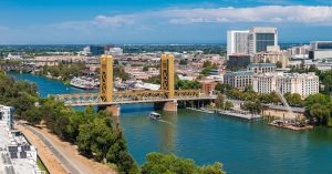Aerial view of Sacramento, California, featuring the golden Tower Bridge over the Sacramento River, a boat, urban buildings, and the State Capitol.