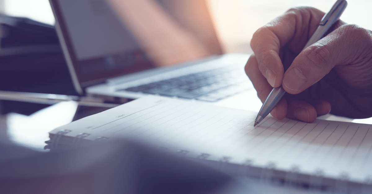 Photo of a hand holding a pen and writing a list next to an open laptop