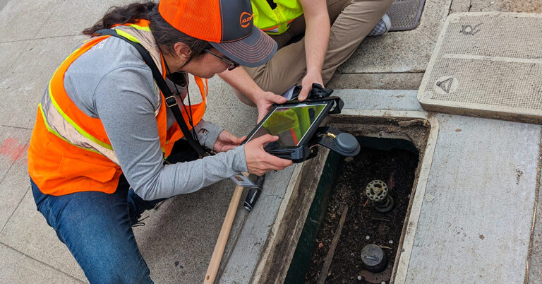 People in safety vests and computer tablet looking into water line under sidewalk