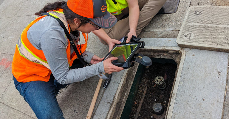 Two people in safety vests holding tablet looking into conduit on sidewalk