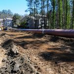 Pipeline above dirt track with heavy equipment and apartment building in background