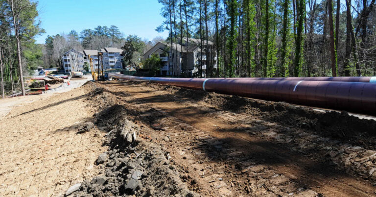 Pipeline above dirt track with heavy equipment and apartment building in background