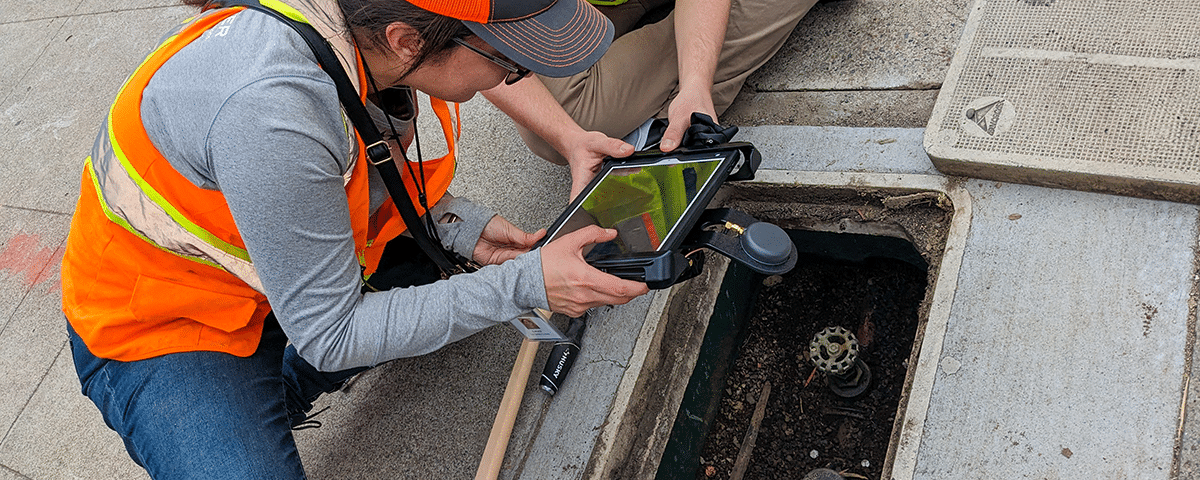 Two people in safety vests looking into hole in ground with computer tablet