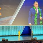 Vic Bianes speaking at PWX conference on large stage with large screen in background