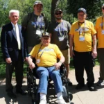 Seven people pose together on a paved path in a green park. One person is seated in a wheelchair wearing a yellow Honor Flight T‑shirt, while six others stand nearby, some also wearing Honor Flight shirts and name badges.