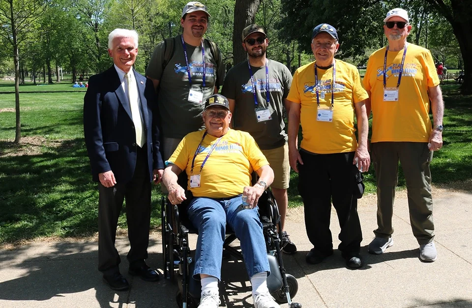 Seven people pose together on a paved path in a green park. One person is seated in a wheelchair wearing a yellow Honor Flight T‑shirt, while six others stand nearby, some also wearing Honor Flight shirts and name badges.
