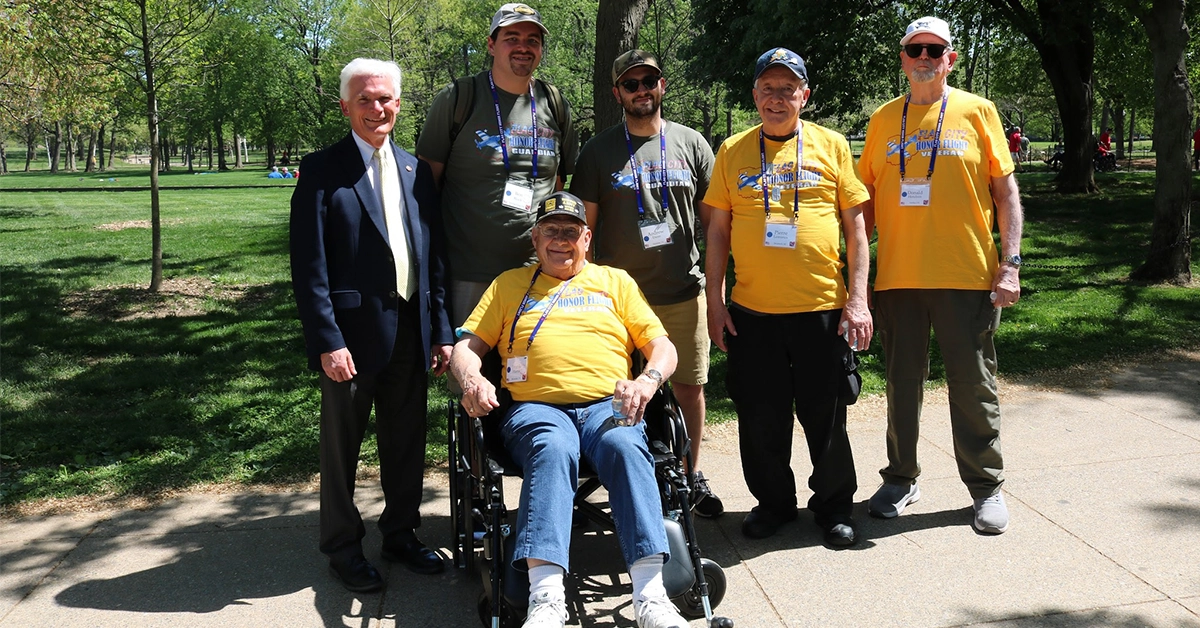 Seven people pose together on a paved path in a green park. One person is seated in a wheelchair wearing a yellow Honor Flight T‑shirt, while six others stand nearby, some also wearing Honor Flight shirts and name badges.