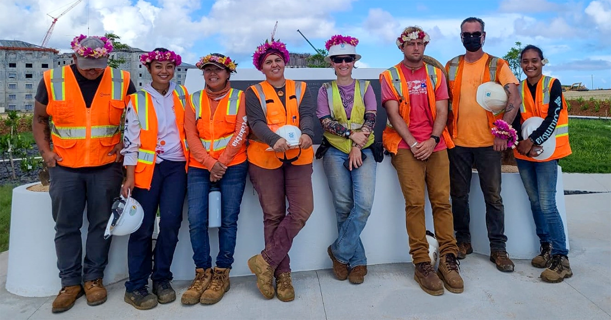 Eight individuals wearing safety gear stand in front of concrete monument with plaque on sunny day