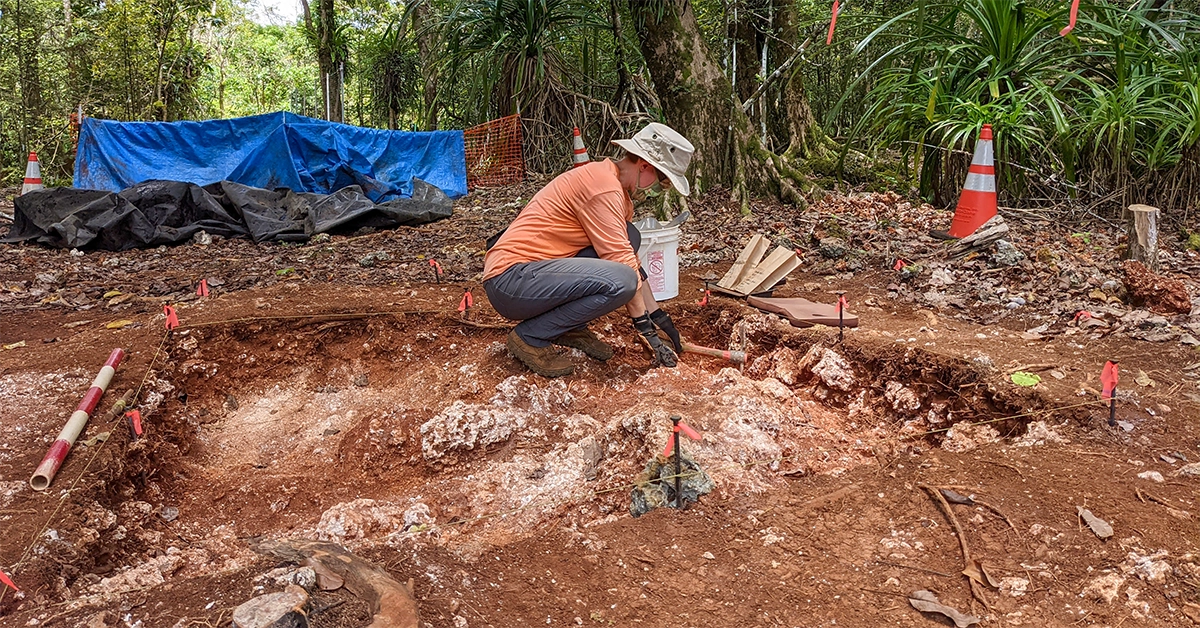 Person wearing orange shirt and brown pants kneels over excavation site with dirt